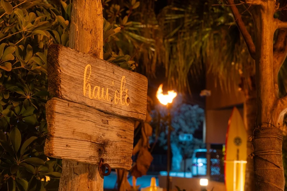 夜のトロピカルな雰囲気の中、木製の看板に「hau'oli」の文字が刻まれています。背景には炎のトーチ、ヤシの木、サーフボードが見え、リゾート地の温かい夜を連想させます。