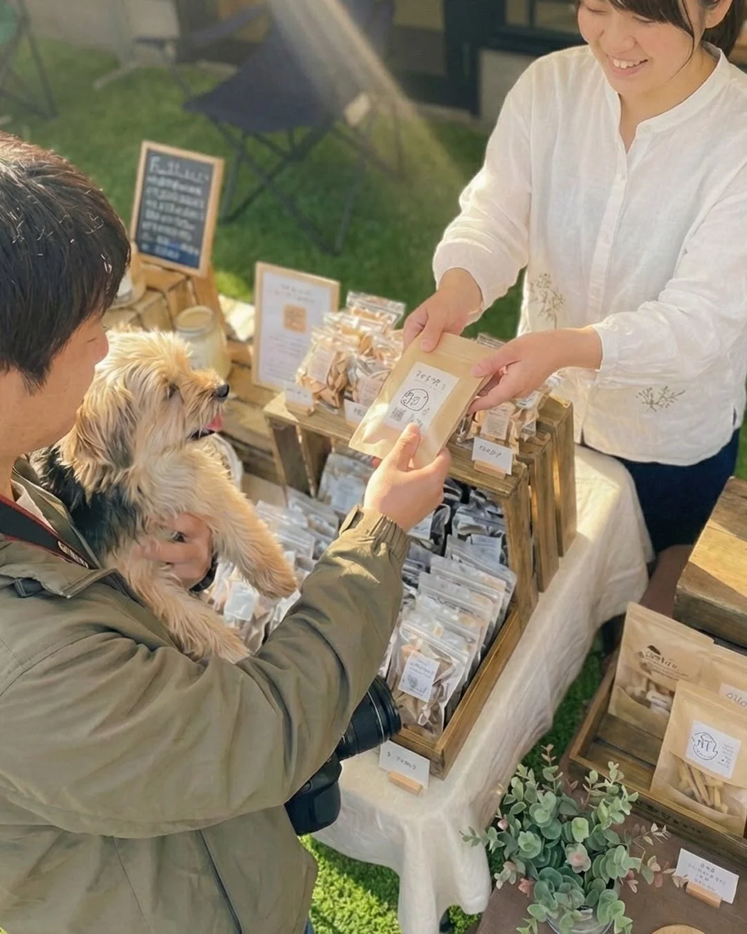 屋外のマルシェで、笑顔の店員が小型犬を抱いた男性客にジャーキーを手渡している