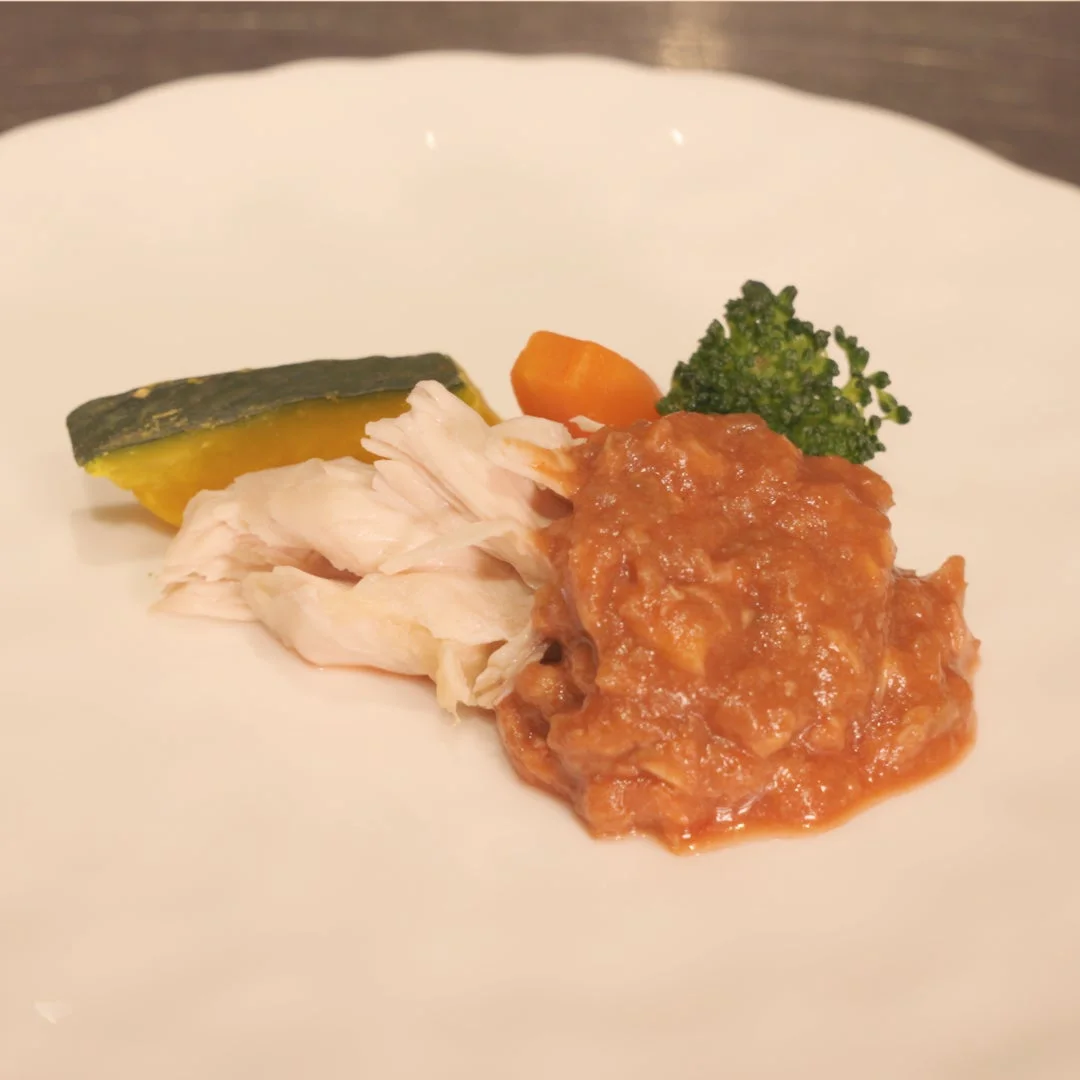白い皿に盛り付けられた、鶏肉のほぐし身とトマトソース、付け合わせにカボチャ、人参、ブロッコリーが添えられた一皿