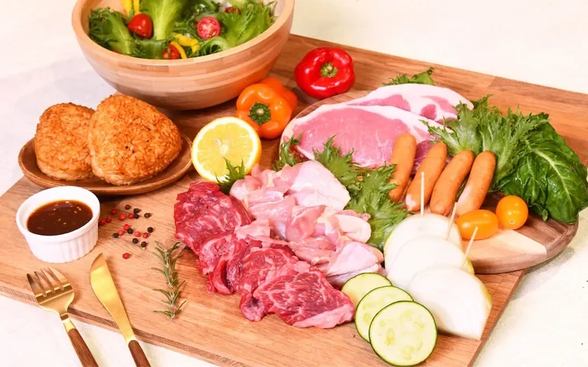 バーベキューや焼肉の準備がされた食材のセットです。牛肉、鶏肉、豚肉、ソーセージなどの肉類と、新鮮な野菜、焼きおにぎり、ソースが木製のボードに美しく盛り付けられています。