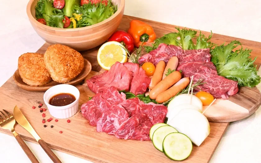焼肉やバーベキューの準備がされた食卓の様子です。木製のカッティングボードには、様々な部位の生肉、ソーセージ、焼きおにぎりが盛り付けられ、レタスやトマト、パプリカなどの新鮮な野菜も添えられています。