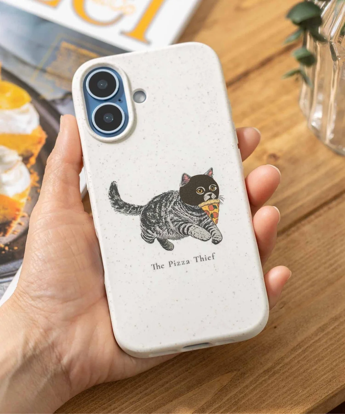 手が白いスマートフォンケースを持っており、ケースにはマスクをかぶってピザをくわえて走る猫のイラストが描かれています。下部には「The Pizza Thief」という文字が見えます。