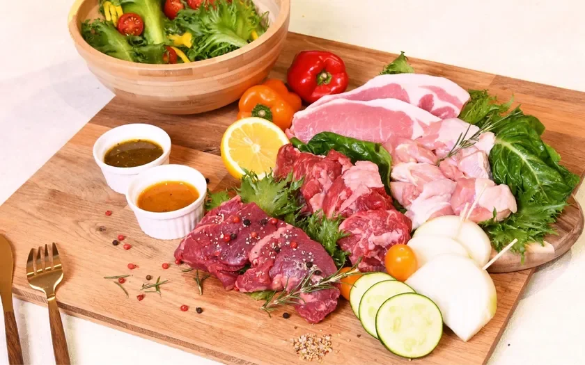木の板の上に、様々な種類の新鮮な生肉（牛肉、豚肉、鶏肉）と、色とりどりの野菜、レモン、2種類のソースが美しく盛り付けられています。奥にはサラダボウルも見え、豪華な食卓の準備風景です。