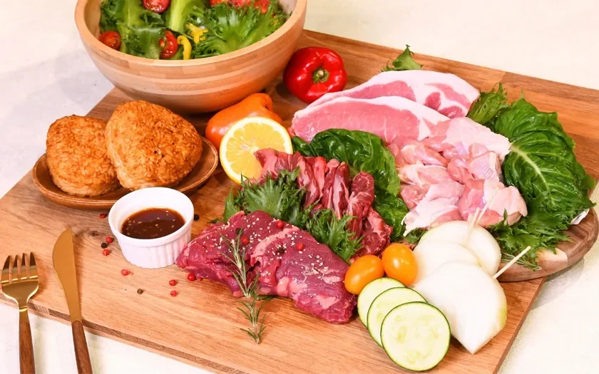 木製ボードに、焼肉や鍋料理にぴったりの新鮮な食材が豊富に並べられています。牛肉、豚肉、鶏肉の生肉、彩り豊かな野菜、焼きおにぎり、サラダが含まれ、食卓の準備風景が伺えます。