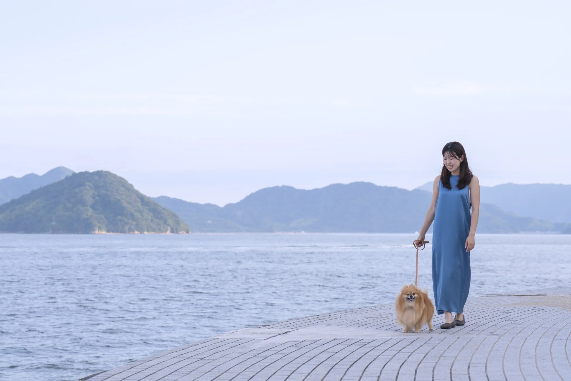 青いロングドレスを着た女性が、ポメラニアンを連れて水辺の舗装された道を散歩しています。背景には穏やかな水面と山々が広がり、晴れた日の気持ちの良い風景です。