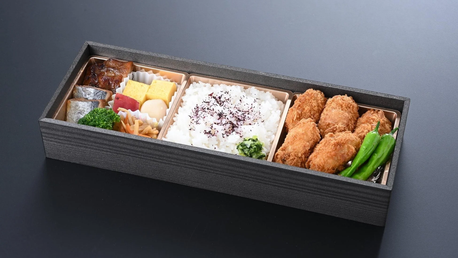木製の箱に詰められた和風の弁当で、白ご飯、焼き魚、揚げ物、たまご焼き、煮物、ししとうなど、彩り豊かなおかずがバランス良く盛り付けられています。